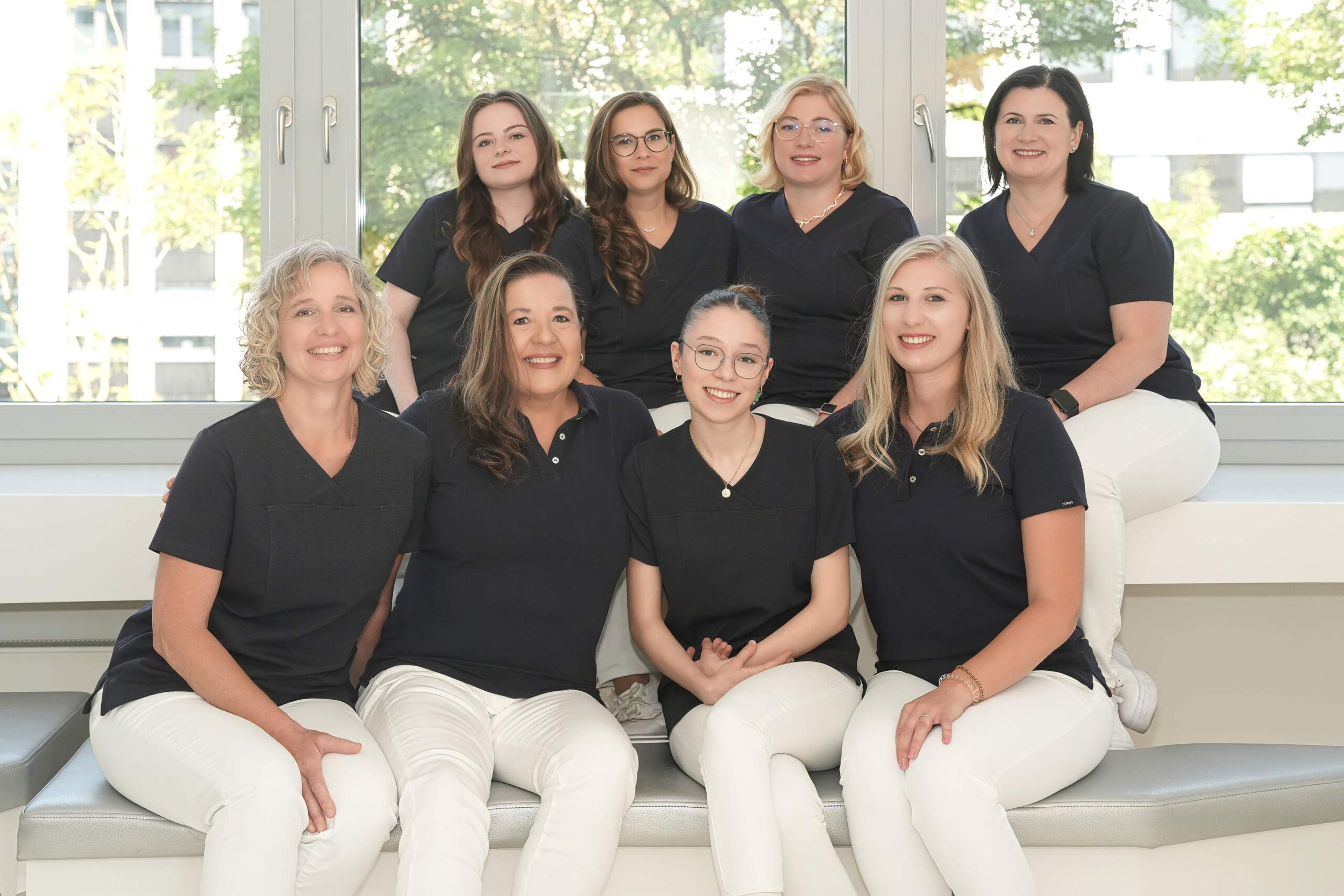 Eight women in black tops and white pants pose together on a white couch by a large window, with greenery outside.