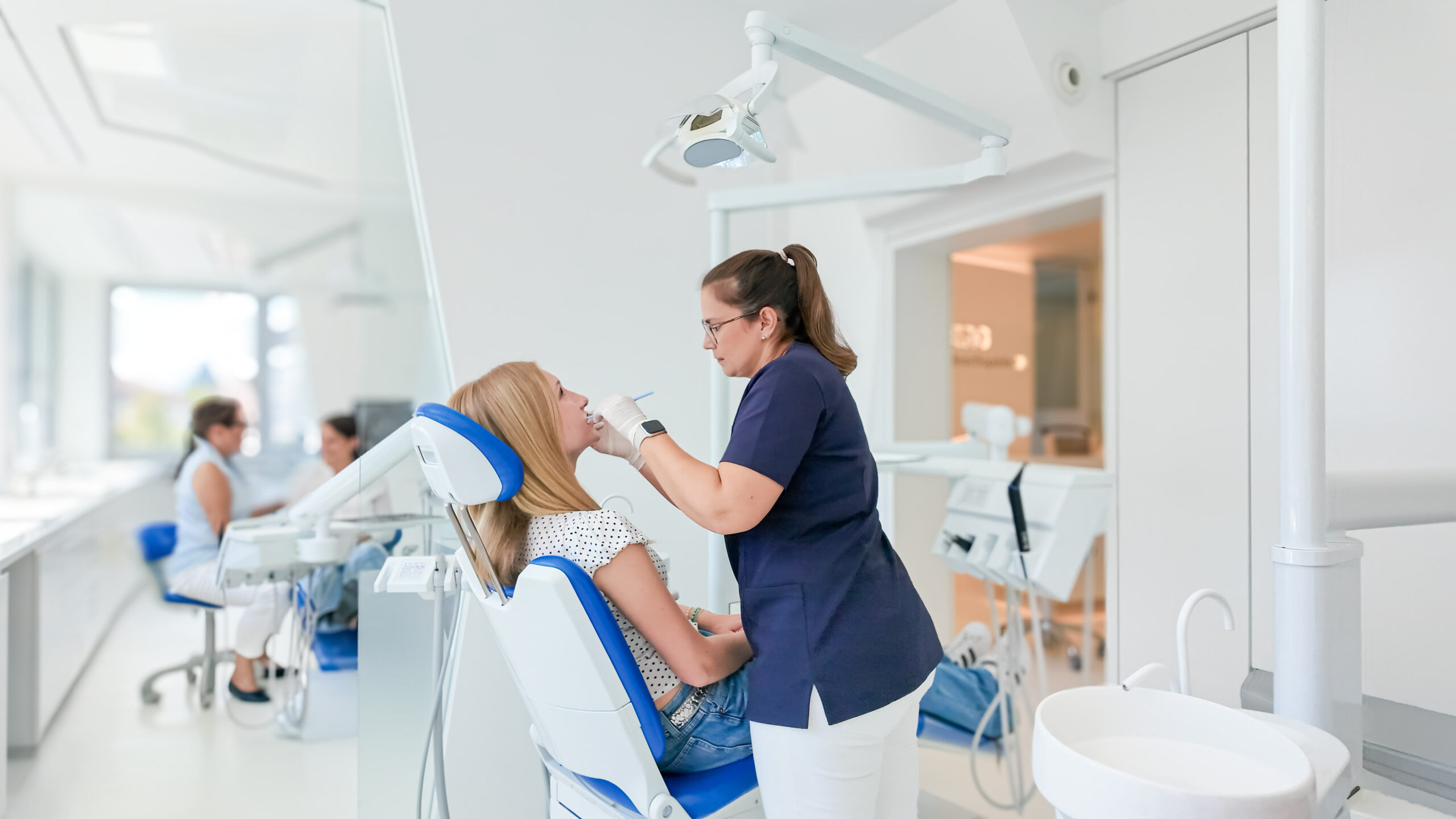 Dentist in navy scrubs treats a woman seated in a blue-and-white dental chair in a bright clinic with dental equipment overhead light nearby.