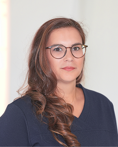 Portrait of a woman with brown hair and glasses, wearing a navy top, standing against a light neutral background.